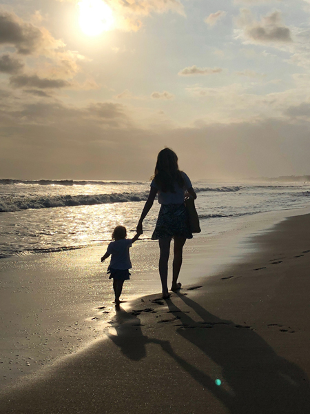 A woman holding a little girl's hand as they walk beside the waves on a beach